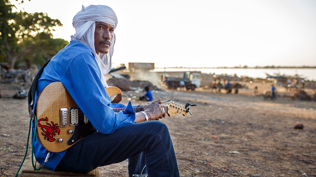 Ahmed Ag Kaedy, wearing a white head covering, sits at the beach holding his electric guitar.